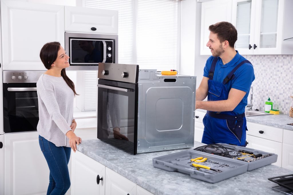 Smiling Repairman Repairing Oven On Kitchen Worktop In Front Of Woman