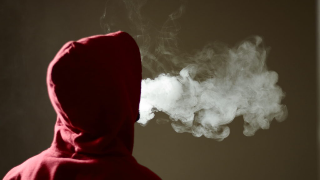 Young male in red hoodie vaping smoking, exhales thick vapor, isolated rear view