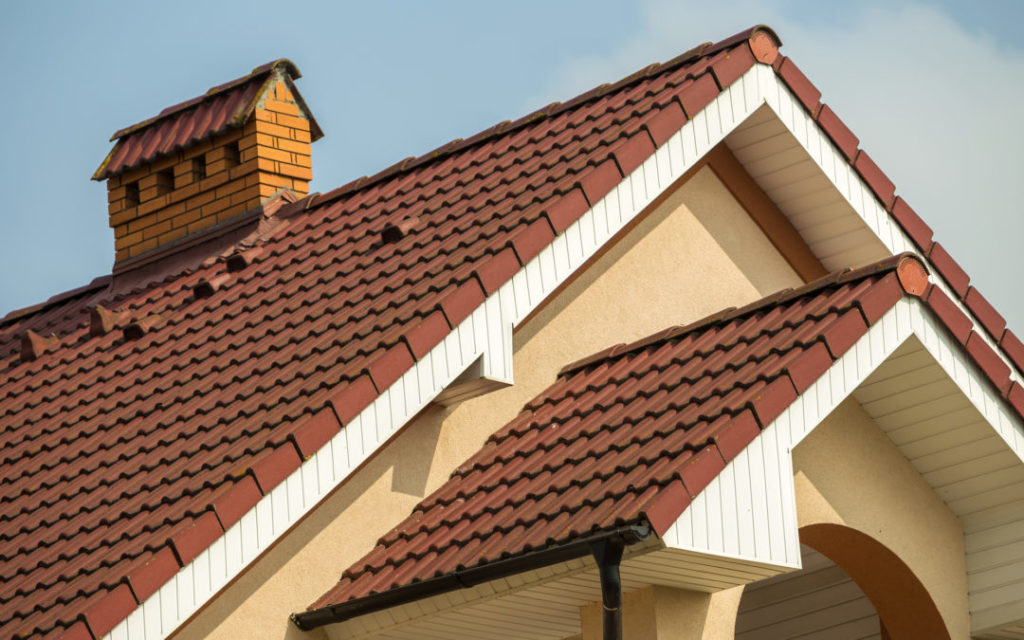 Top detail of big modern expensive residential cottage with steep shingled roof, brick chimney, stucco walls on blue sky background. Construction, professional roofing and investment concept.