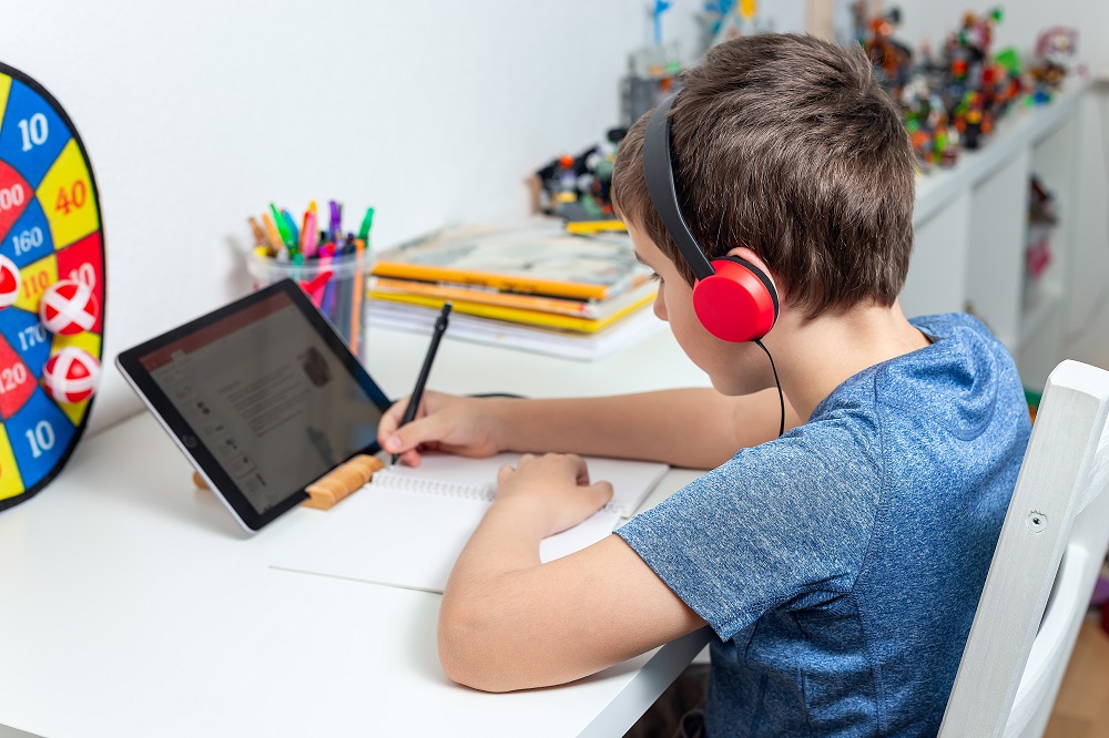 Eight years old boy behind the table study at home using a tablet and headphones, digital education, homeschooling at isolation