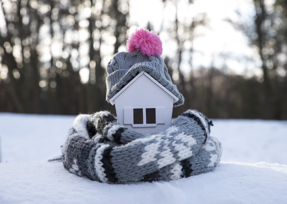 house in winter – heating system concept and cold snowy weather with model of a house wearing a knitted cap
