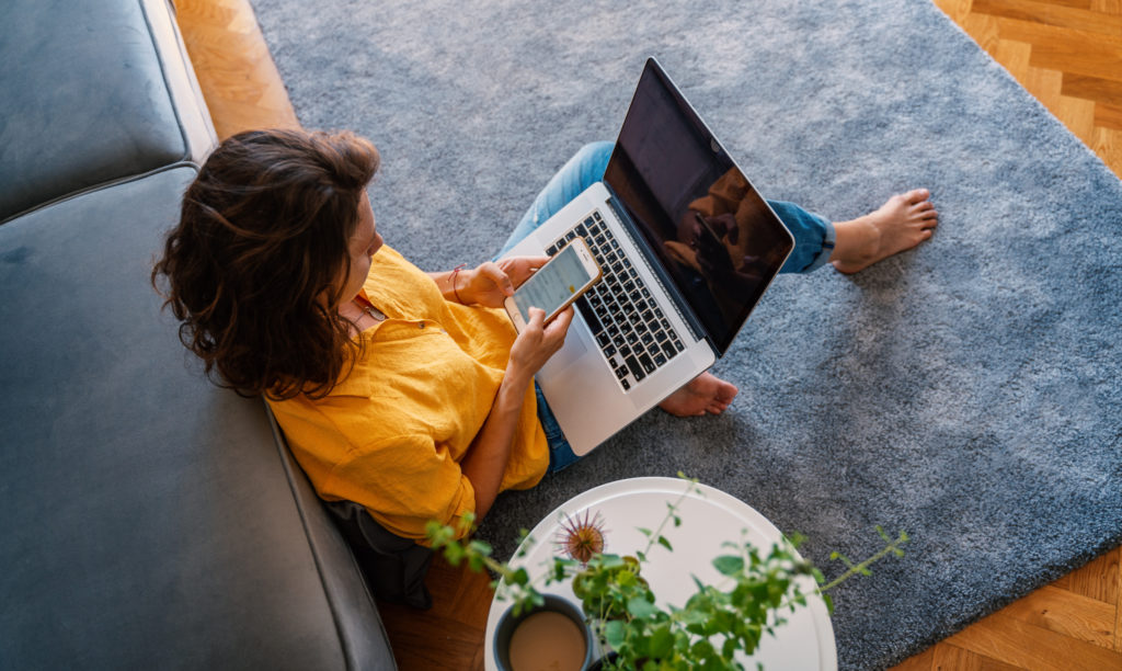 young girl in a yellow shirt woman working on a laptop at home using a smartphone in the living room on the carpet, distant work and education