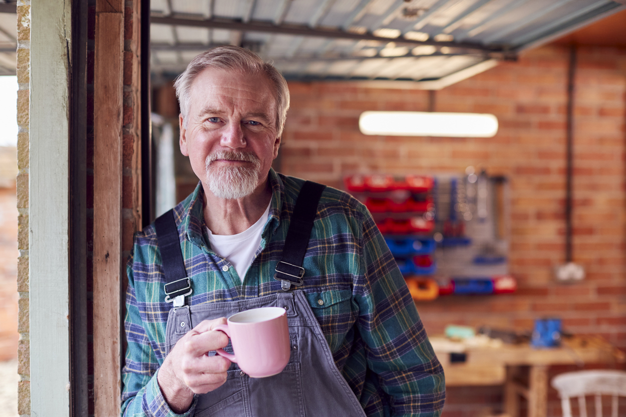 Portrait Of Senior Male Wearing Overalls In Garage Workshop With Hot Drink